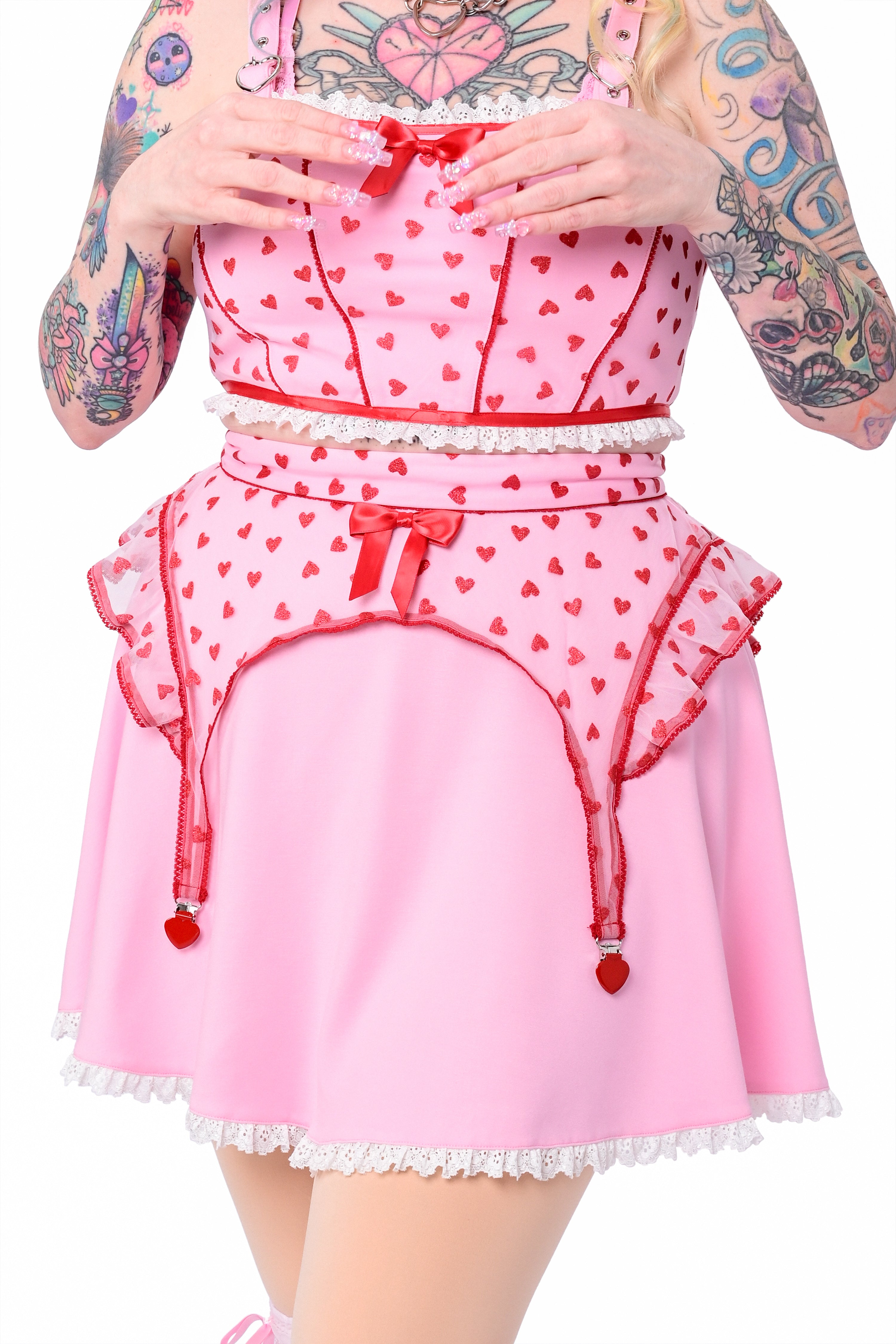 A model wearing a pink skater skirt with a lace hem and glittery heart details. The skirt has red accents and a zip-up back.