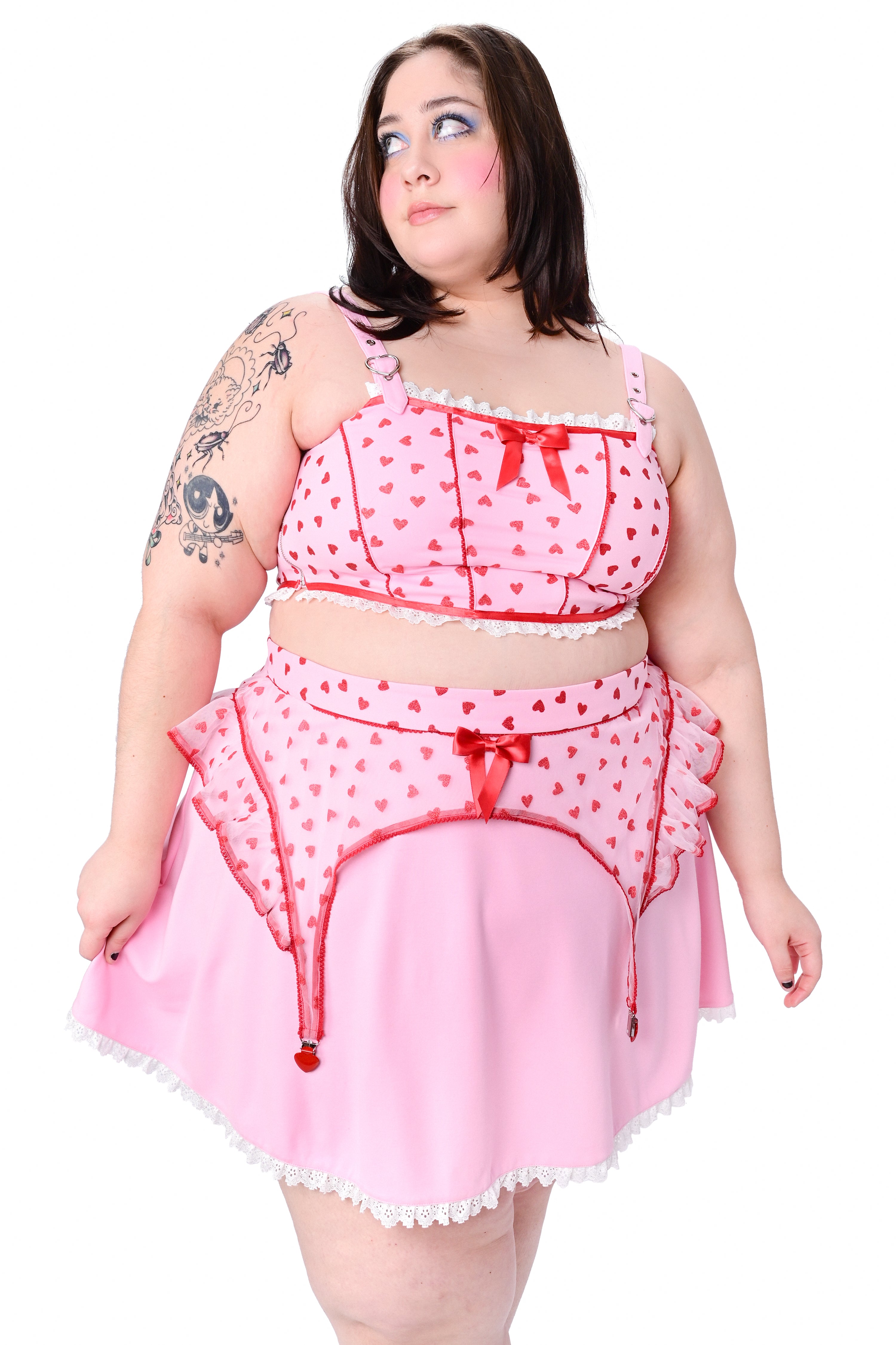 A person wearing a pink corset style crop top with a red mesh overlay, adorned with glittery hearts and a dainty pink bow at the neckline