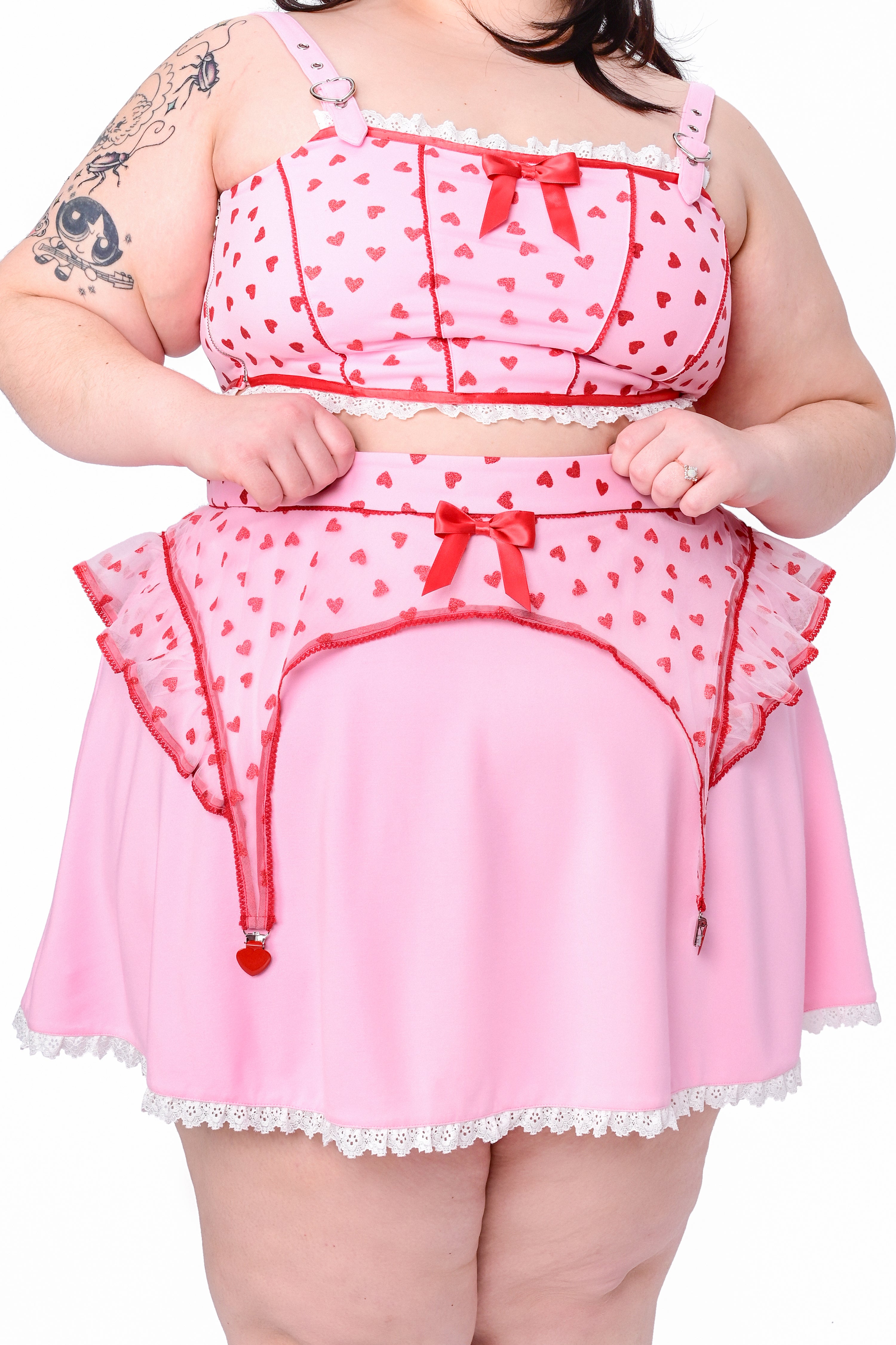 A model wearing a pink skater skirt with a lace hem and glittery heart details. The skirt has red accents and a zip-up back.