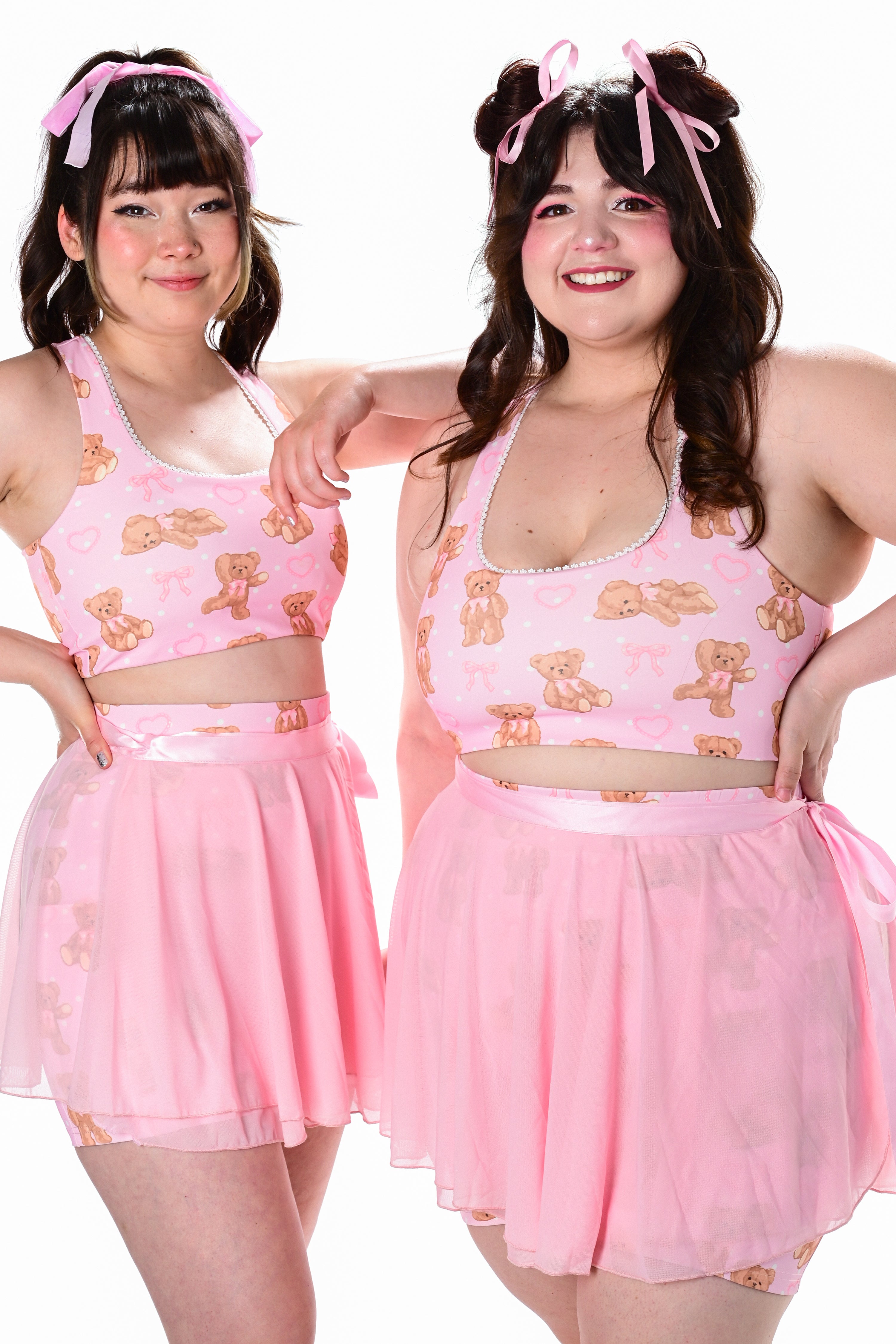 Two models wearing pink activewear outfits with a teddy bear print. The outfits include biker shorts and a wrap skirt.