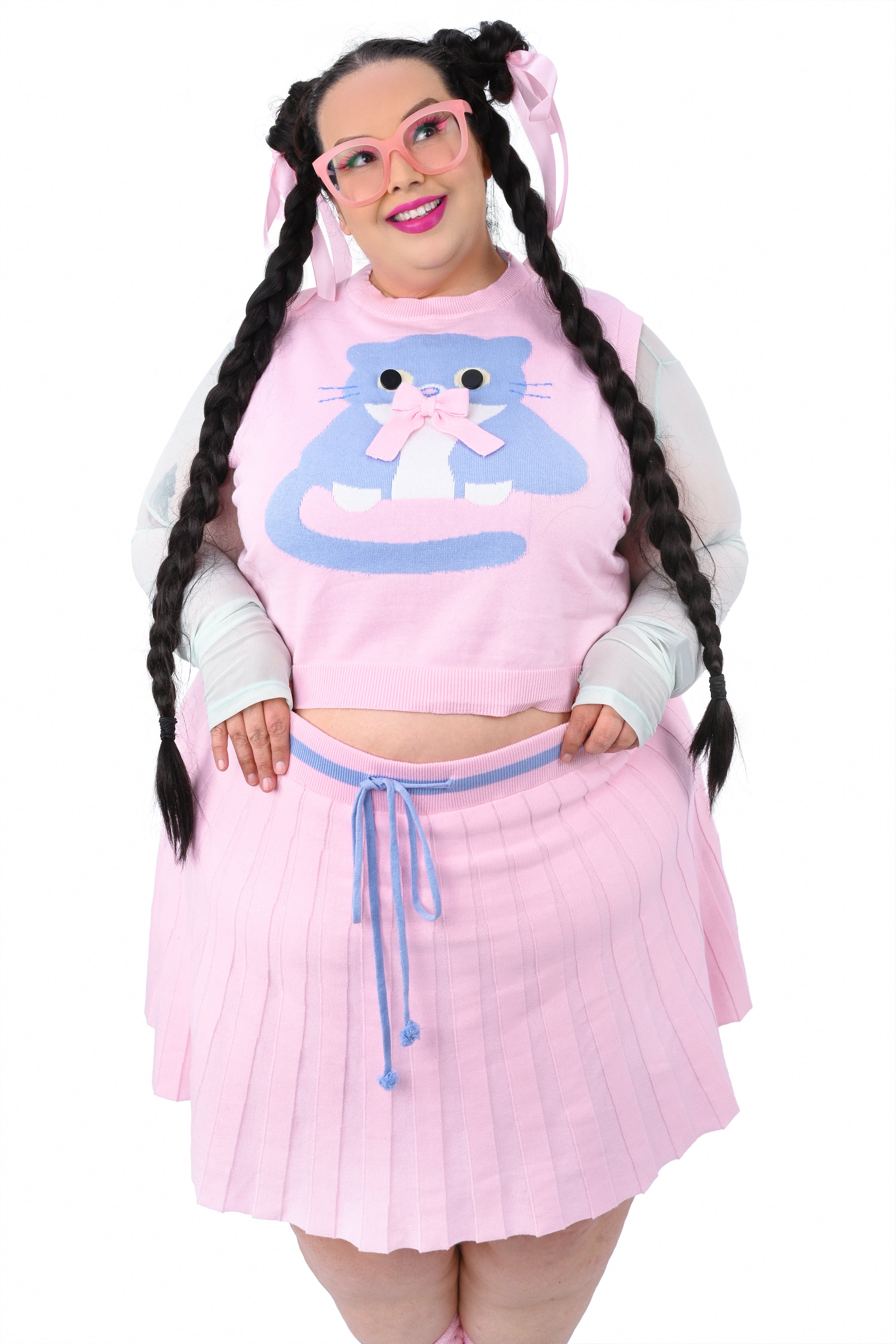 A person wearing a pink knit sweater vest with a large button-eyed cat design, paired with a matching pleated skort