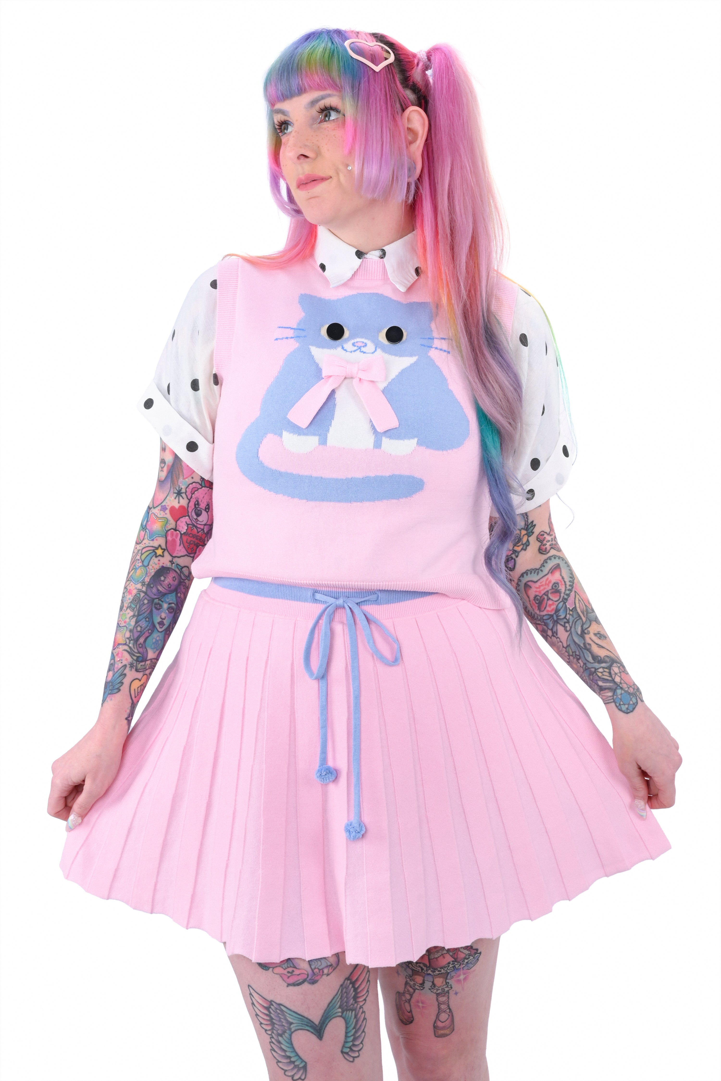 A person wearing a pink knit sweater vest with a large button-eyed cat design, paired with a pleated skort and a polka dot shirt.