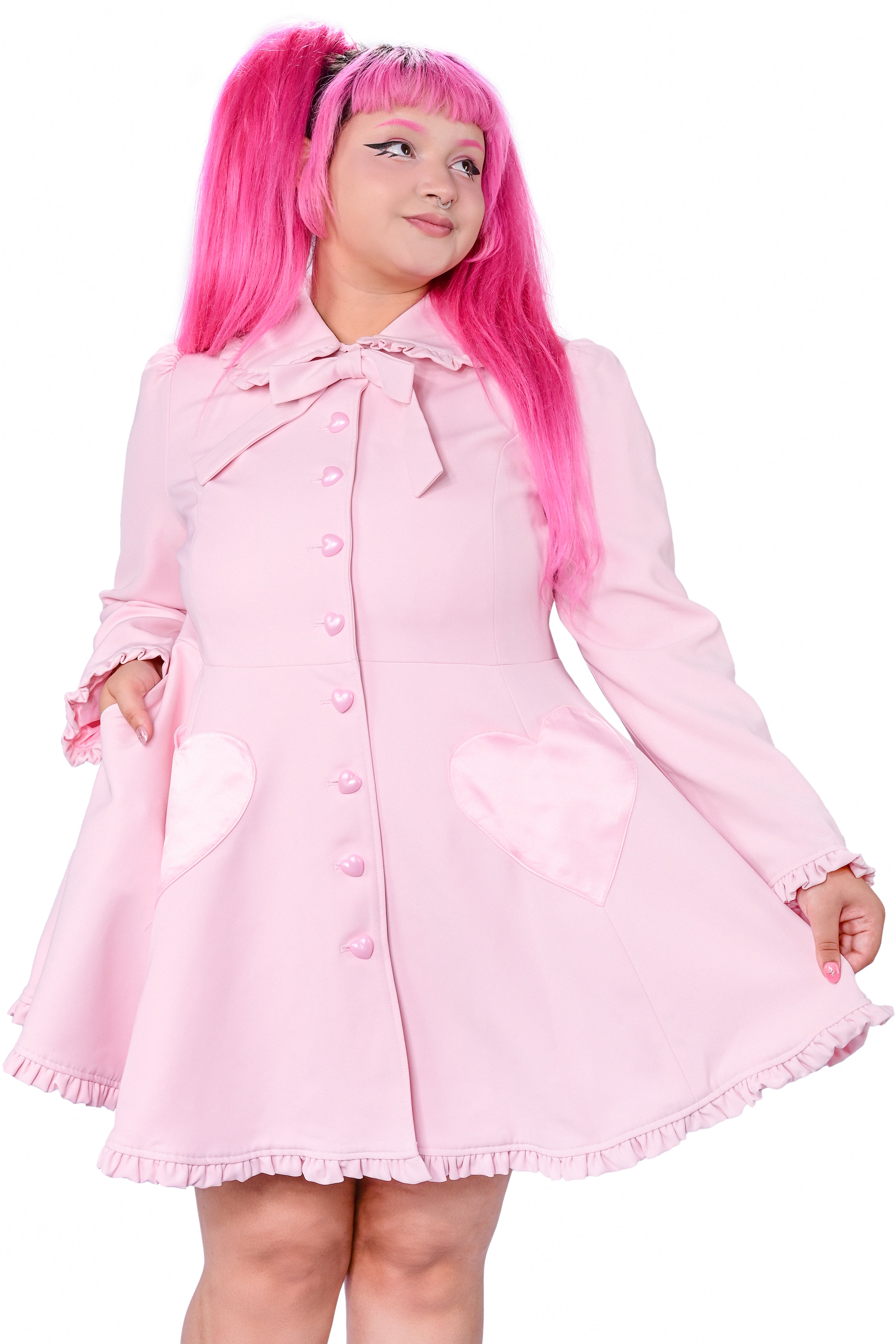 A person modeling a pink coat with detachable bunny ears and frilly details at the collar and hem. The coat features heart-shaped pockets and buttons, and is fitted at the bodice.