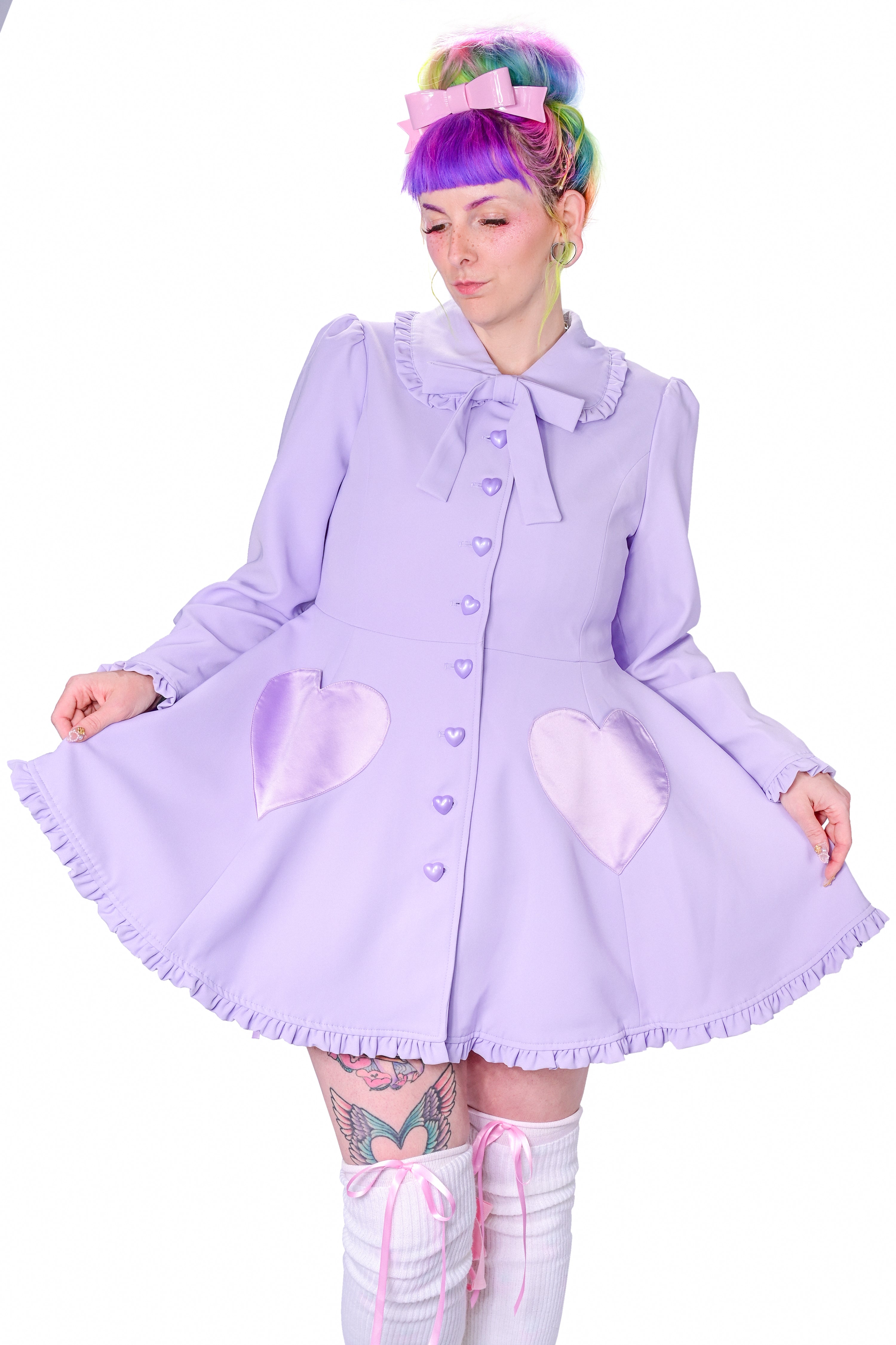 A person modeling a lavender coat with detachable bunny ears and frilly details at the collar and hem. The coat features heart-shaped pockets and buttons, and is fitted at the bodice.