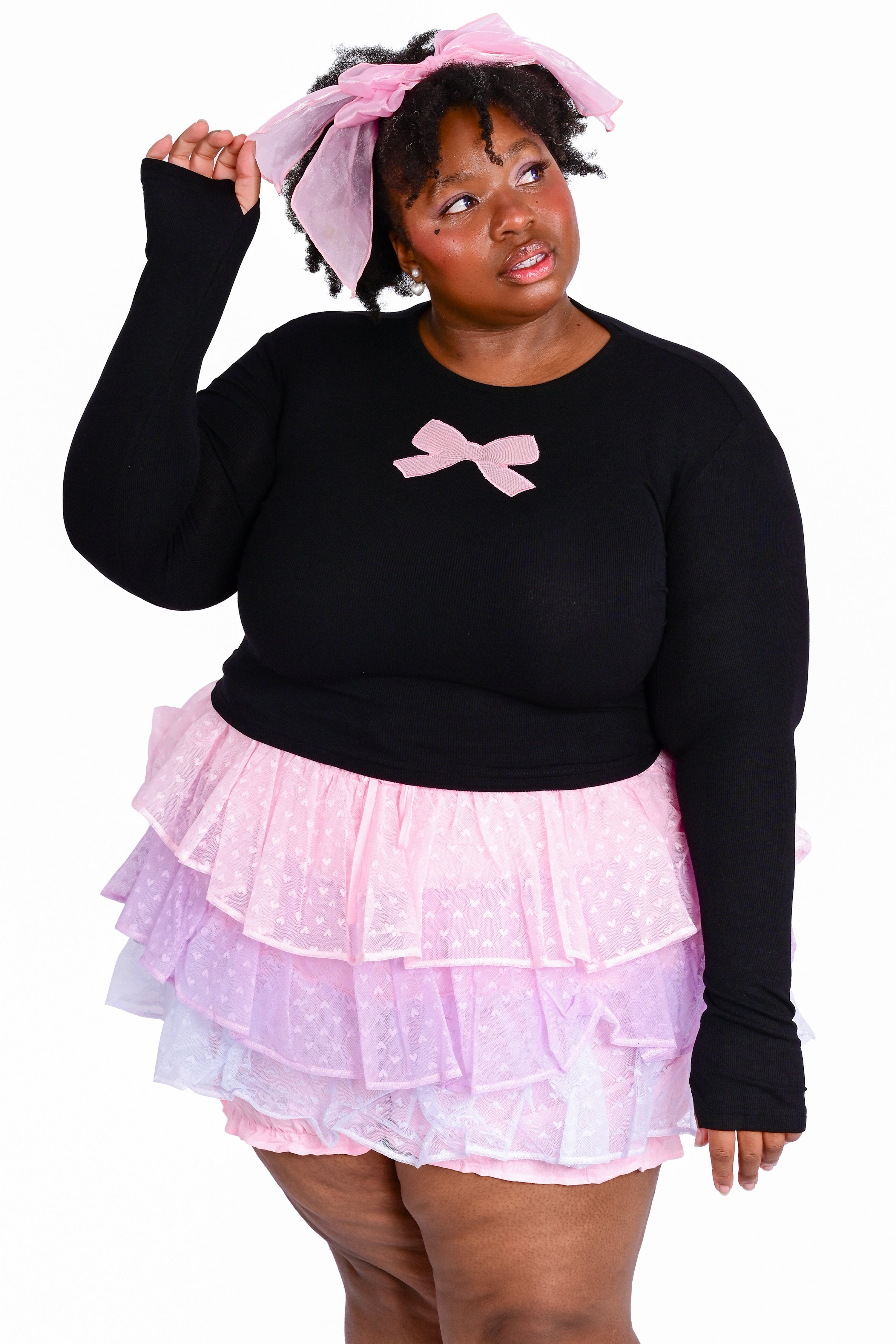 A person wearing a black long sleeve top with a pink ribbon bow cutout on the front and a tiered ruffle skort