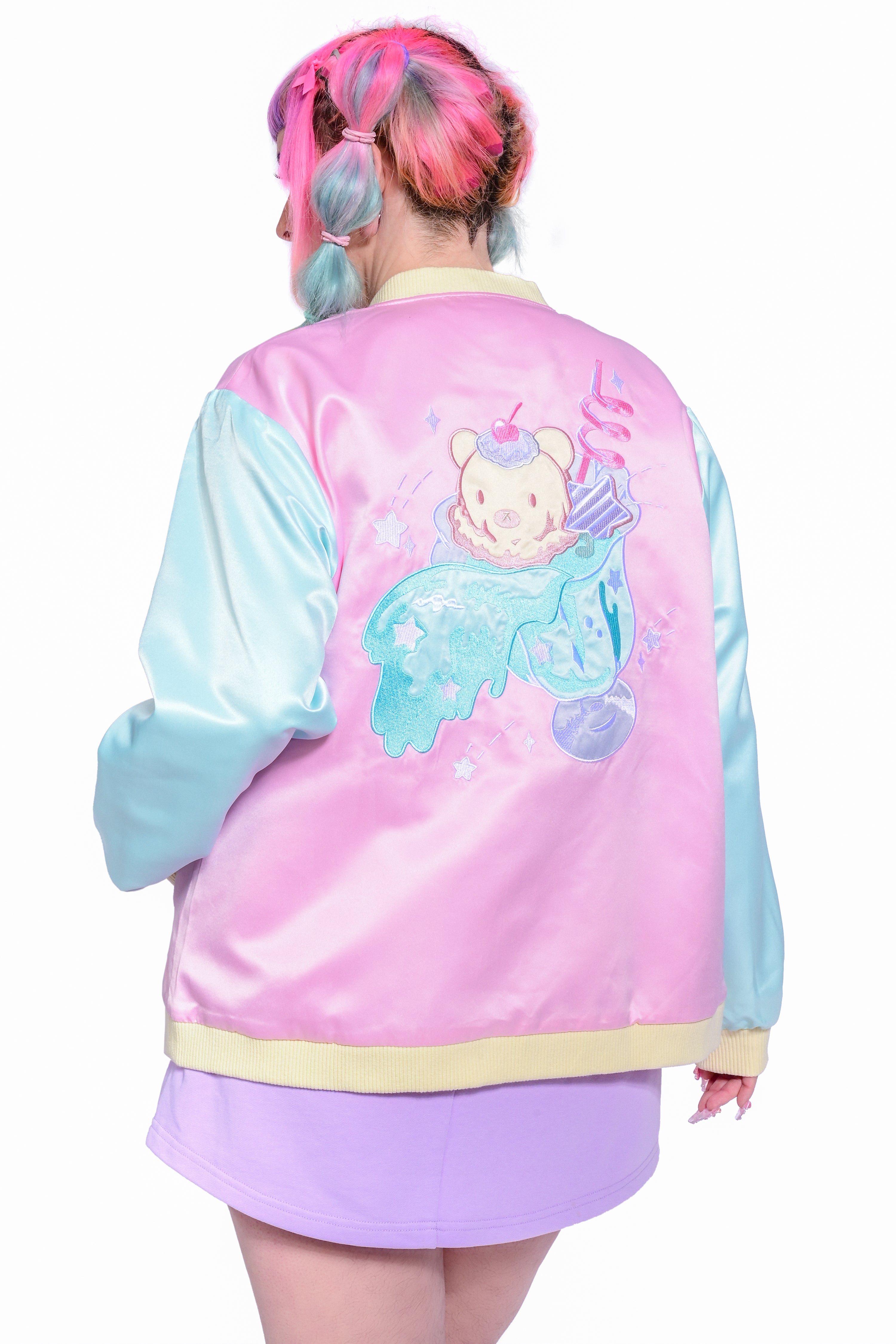 A person wearing an embroidered satin color blocked varsity jacket in pink, blue, and yellow. The back features an applique in the image of a soda float.