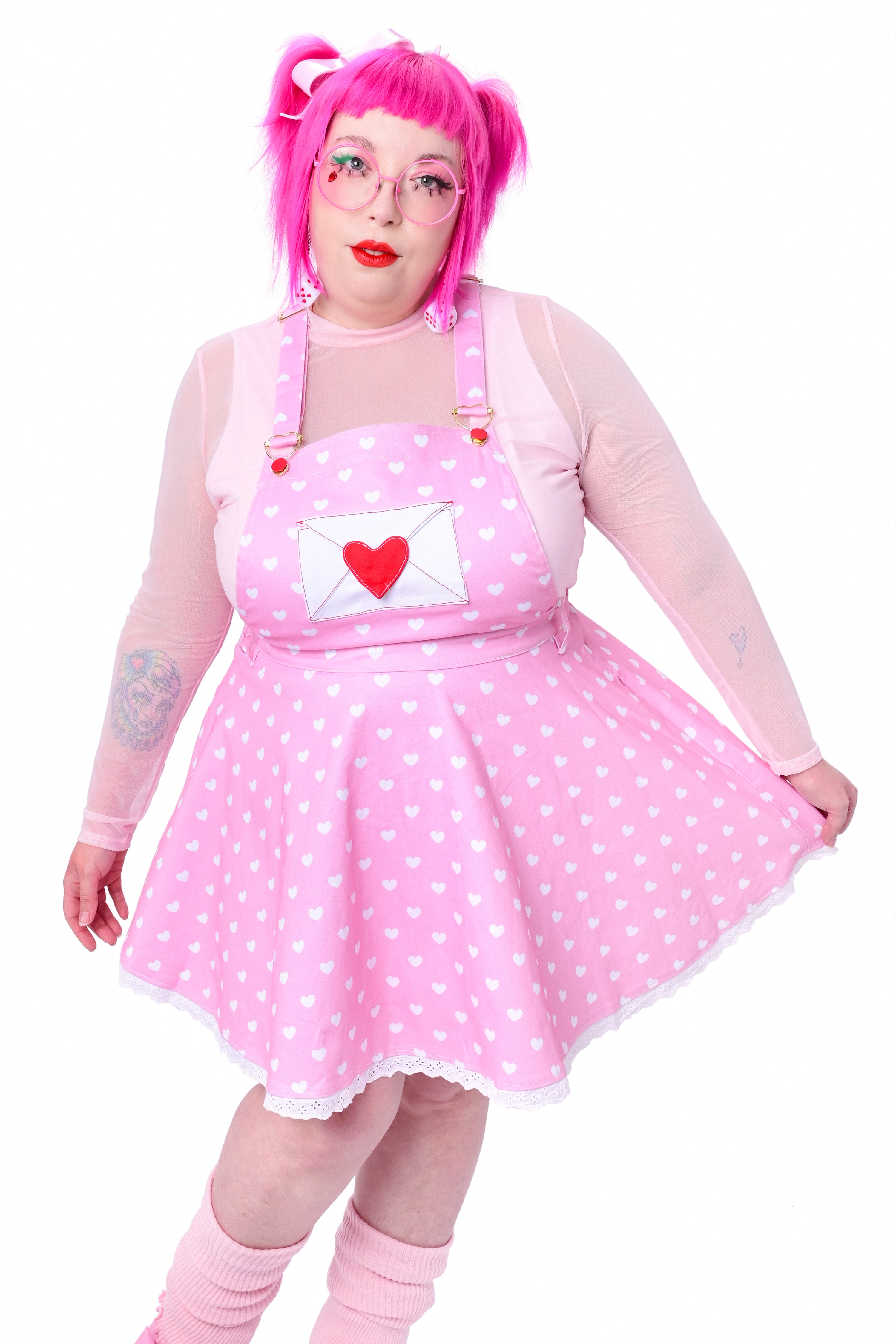 A person wearing a pink heart-printed pinafore dress with red buttons and heart-shaped hardware on the straps.
