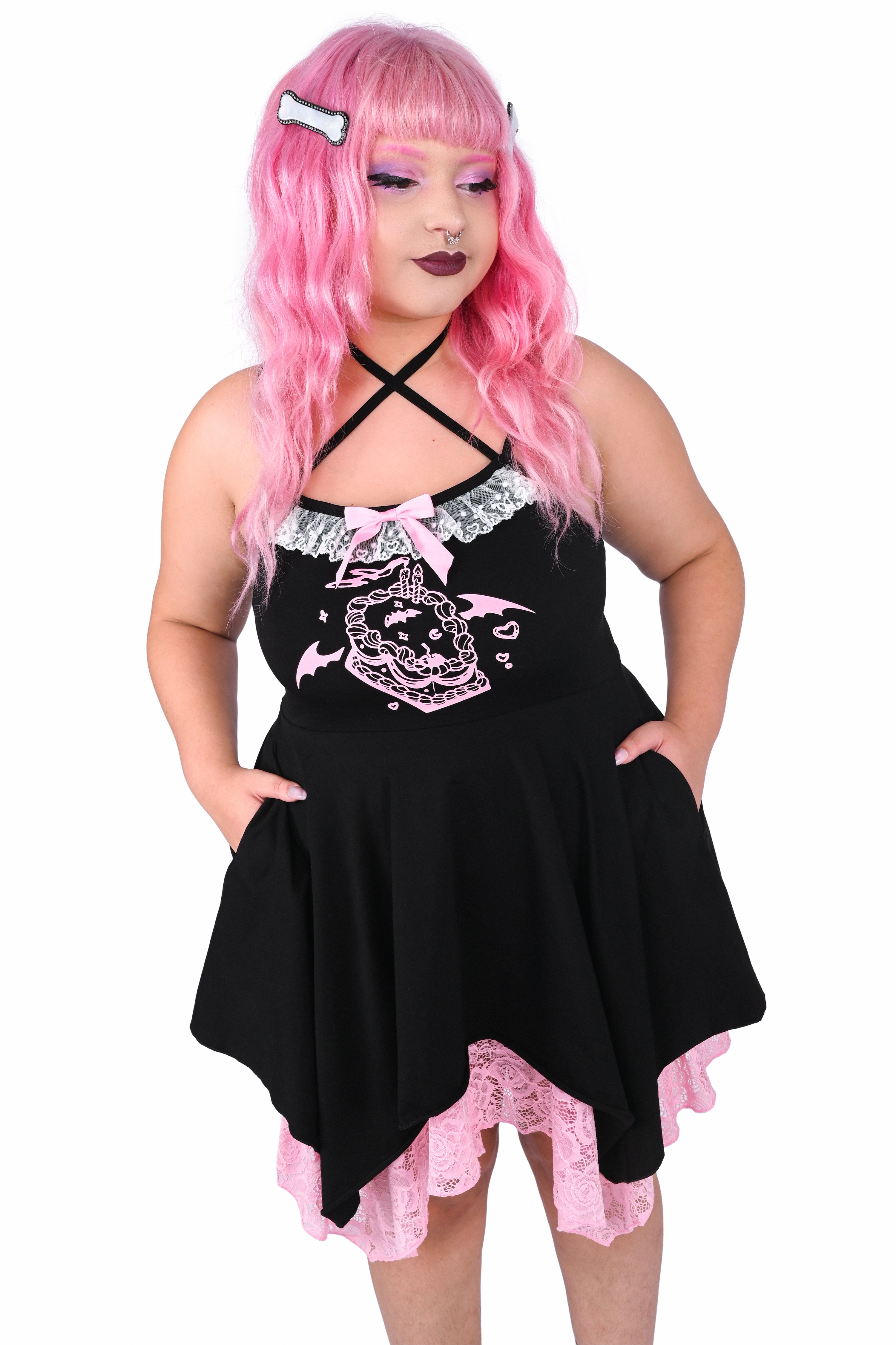 Model wearing a black dress with a cake graphic and pink and white lace details. The model's hands are placed in the dress's pockets.
