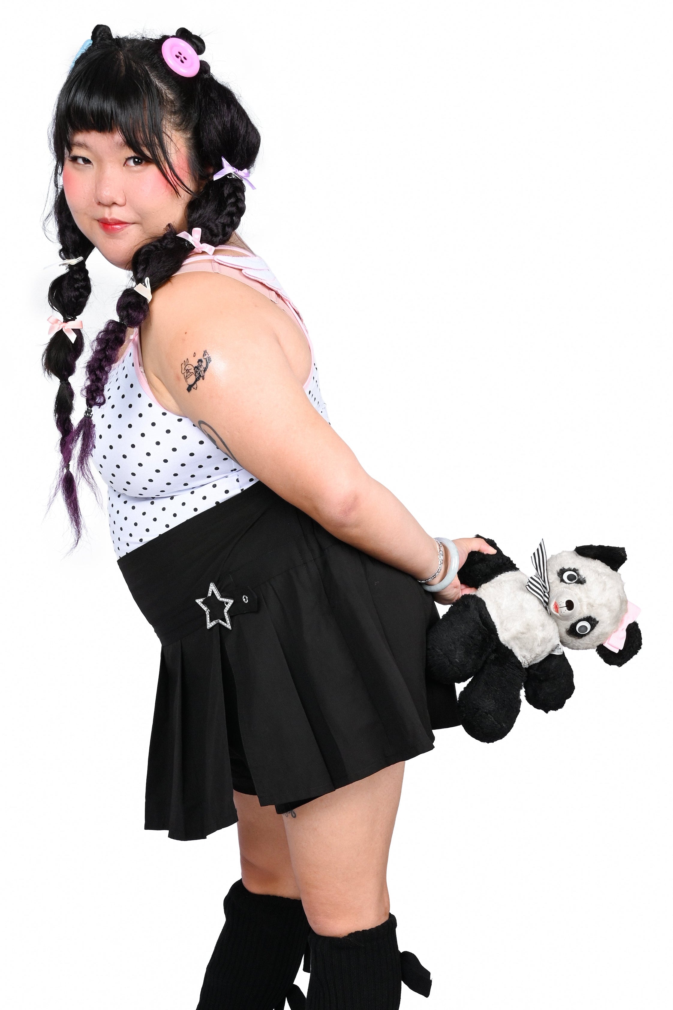 A person modeling a goth-style outfit consisting of a white sleeveless top with black polka dots, a black pleated mini skirt with a silver star, black knee-high leg warmers, and black shoes, while holding a small black-and-white plush toy against a plain white background.