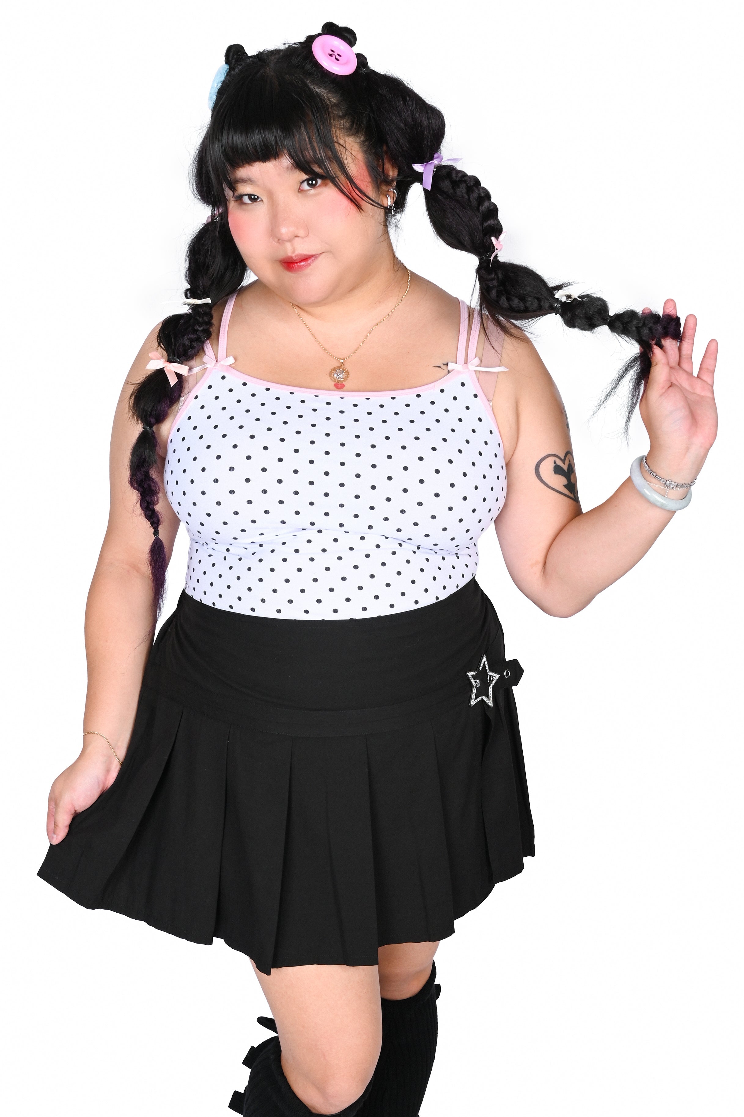 A person modeling a cute outfit consisting of a white tank top with black polka dots, a black pleated mini skirt with a silver star buckle, black leg warmers, and black shoes, against a plain white background.