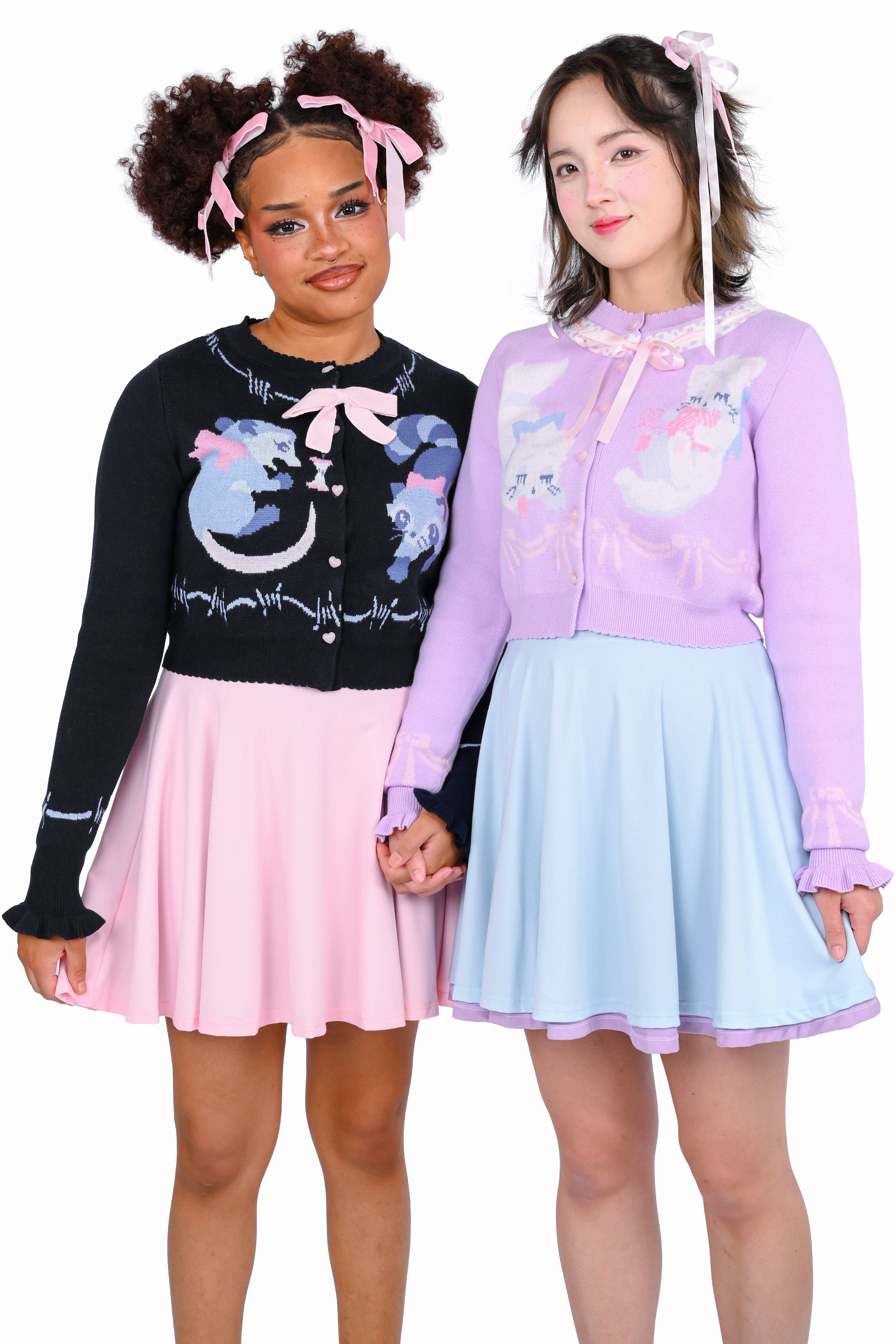 Two models, one wearing a navy cardigan with a raccoon and possum graphic, and the other wearing a lavender cardigan with cat graphics