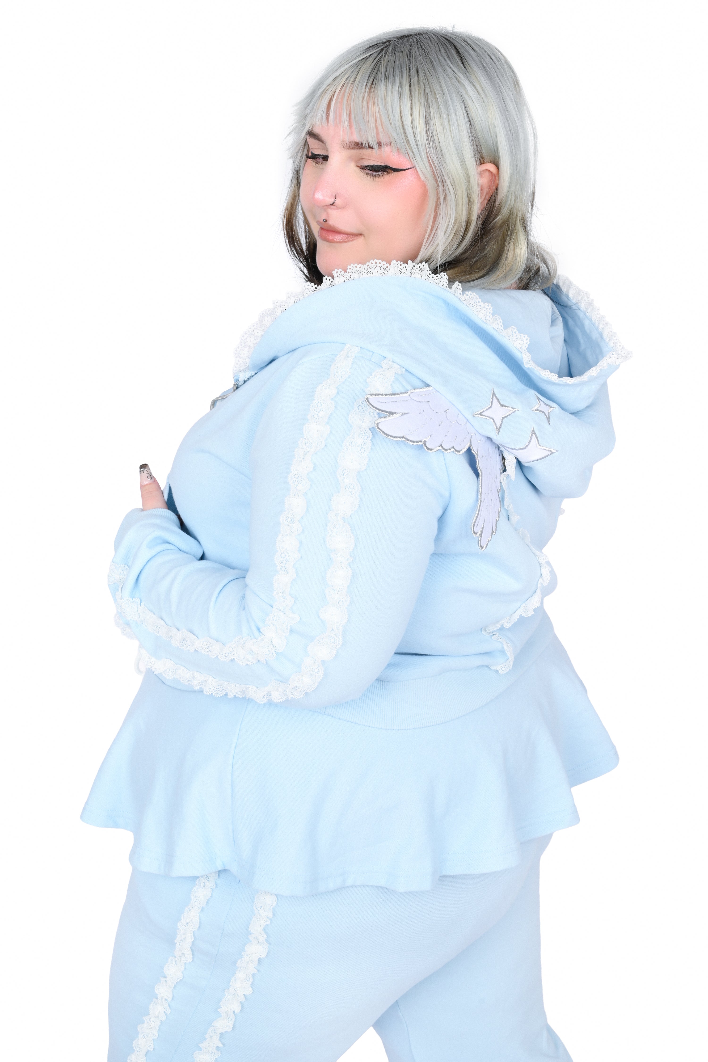 A model with silver hair wearing a light blue angel-themed track set, featuring a hooded jacket with white lace trim, angel wings on the shoulder, and matching ruffled pants, shot against a plain white background.