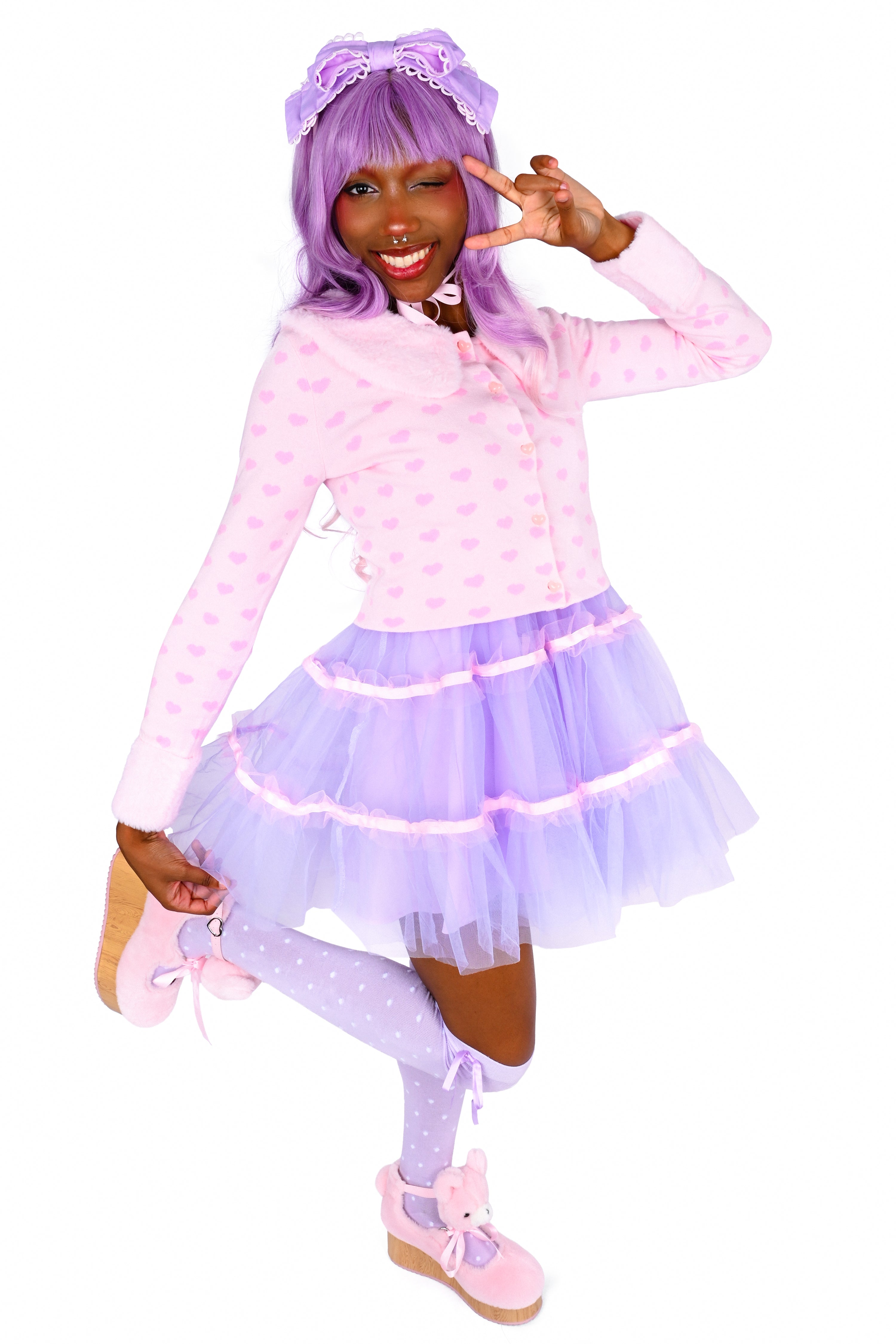 A person modeling a lavender and pink outfit, featuring a polka-dot pink long-sleeve top, a tiered purple tulle skirt with pink trim, matching purple polka-dot tights, a purple wig with a bow, and pink shoes, posed against a solid white background.