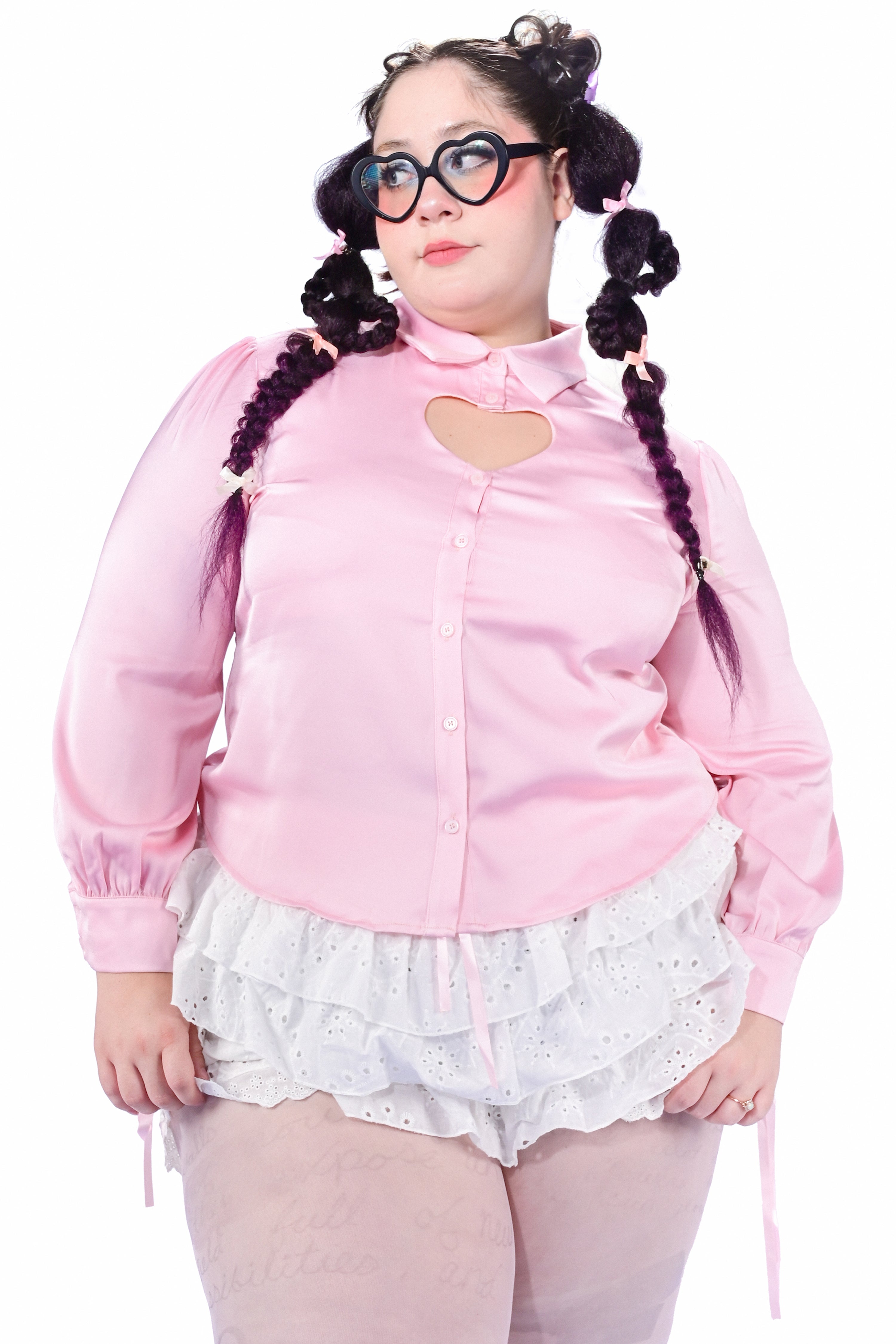 A model wearing a pink button-up blouse with a heart cutout at the chest, paired with white lace shorts.