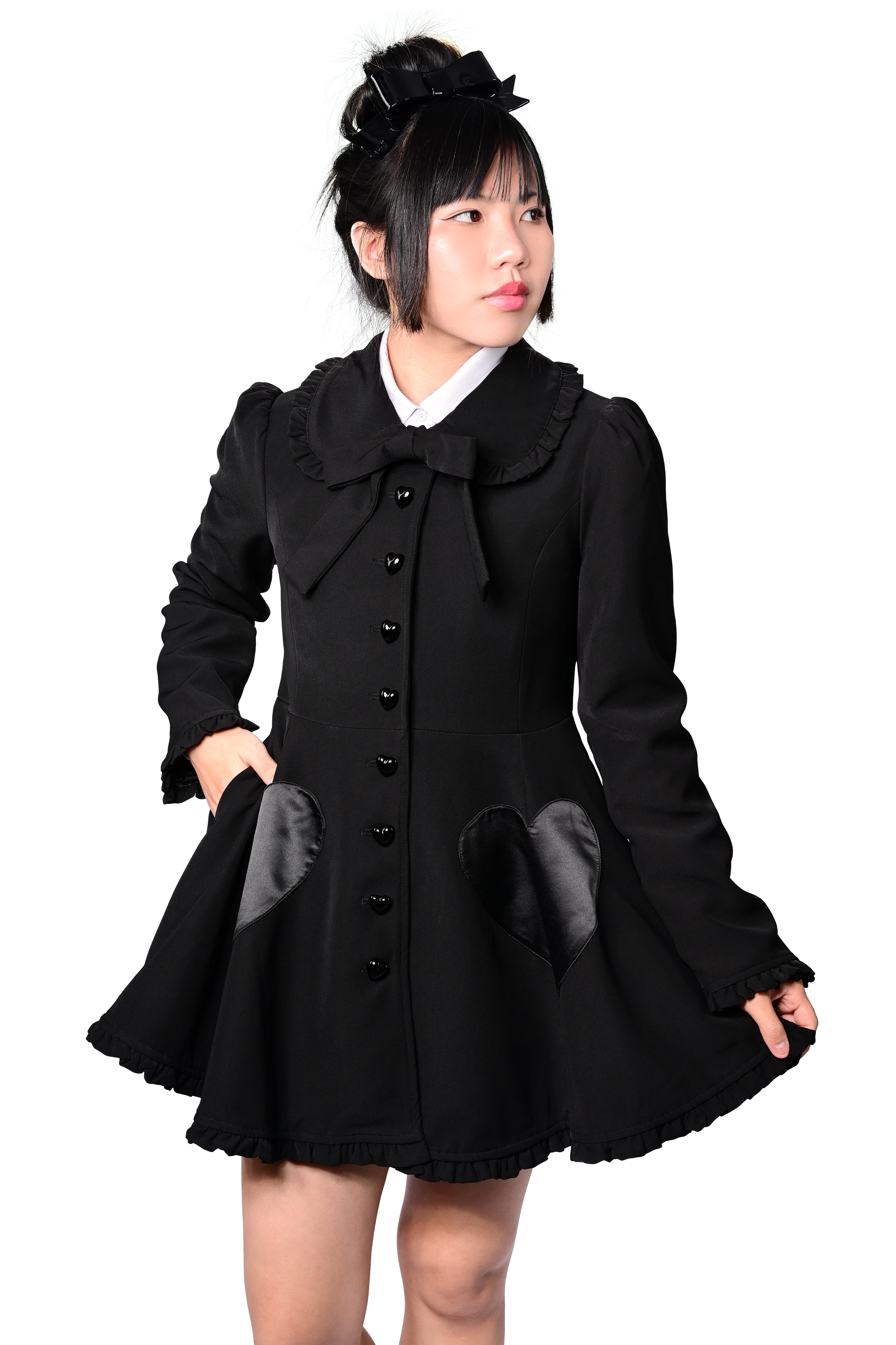A person modeling a black coat with detachable cat ears and frilly details at the collar and hem. The coat features heart-shaped pockets and buttons, and is fitted at the bodice.