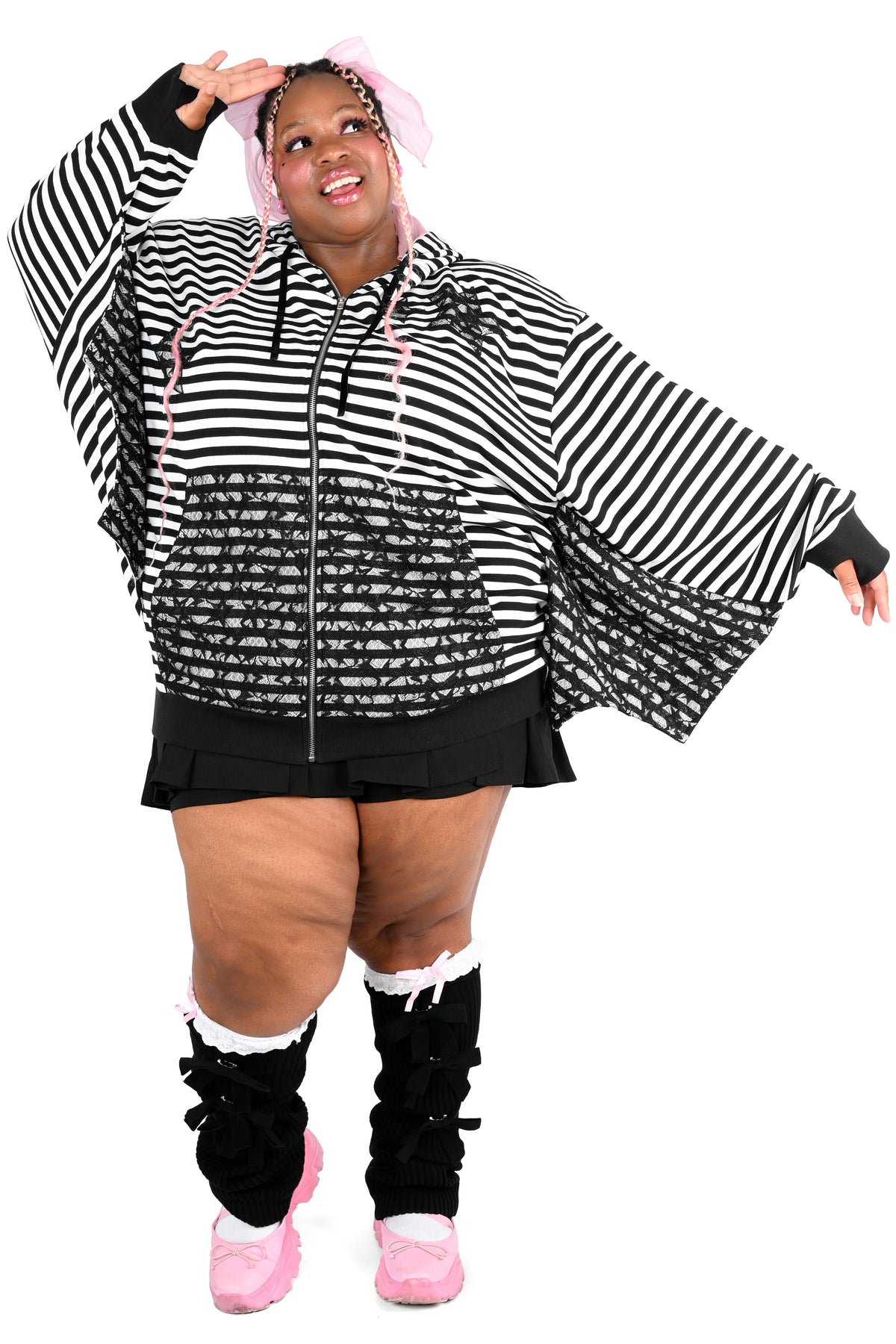 A plus-size model wearing a black and white striped bat wing hoodie, black pleated mini skirt, black leg warmers, and pink sneakers, on a solid white background.