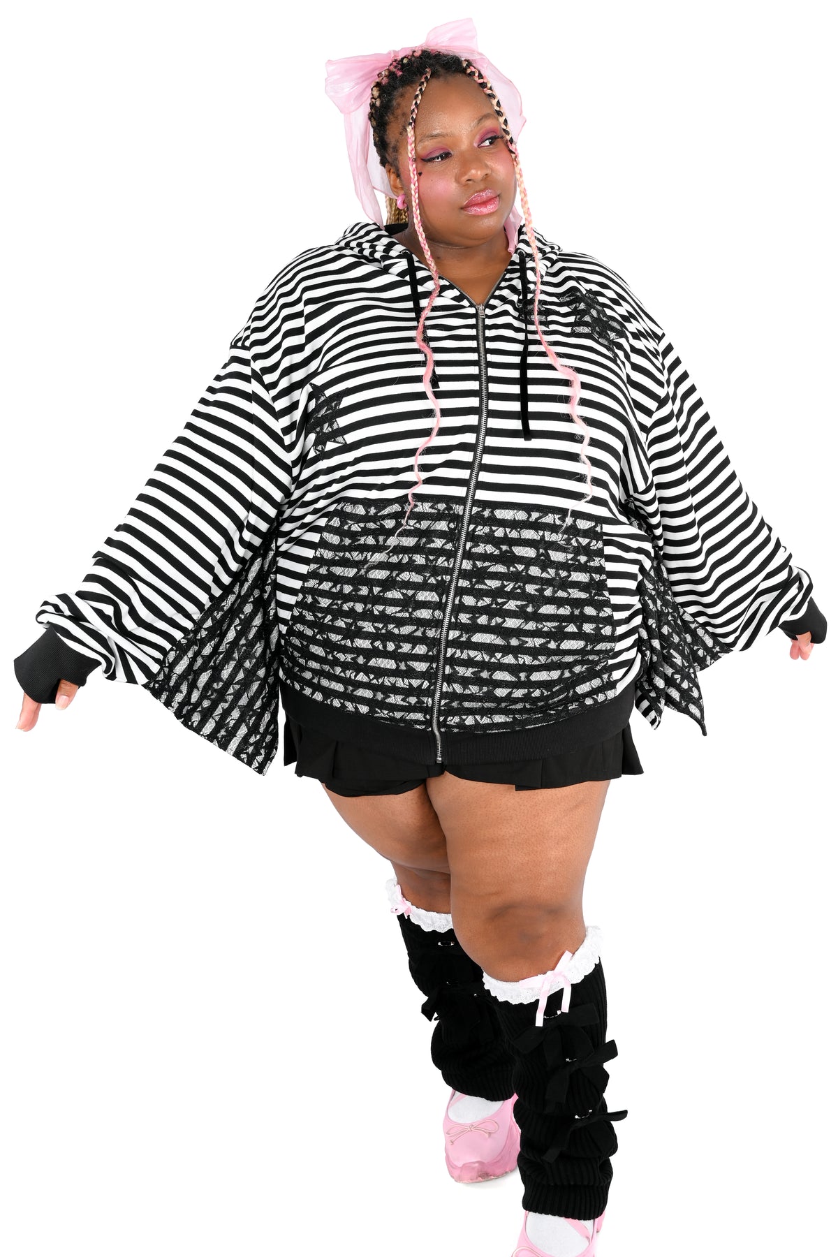 A plus-size model wearing a black and white striped zip-up hoodie with a black lace overlay on the pockets and on the chest- The chest overlays are in the shape of stars. There are 2 stars on her left upper chest and one star on the lower right chest. She is wearing black shorts, black leg warmers with white ruffle trim, and pink sneakers, set against a plain white background.