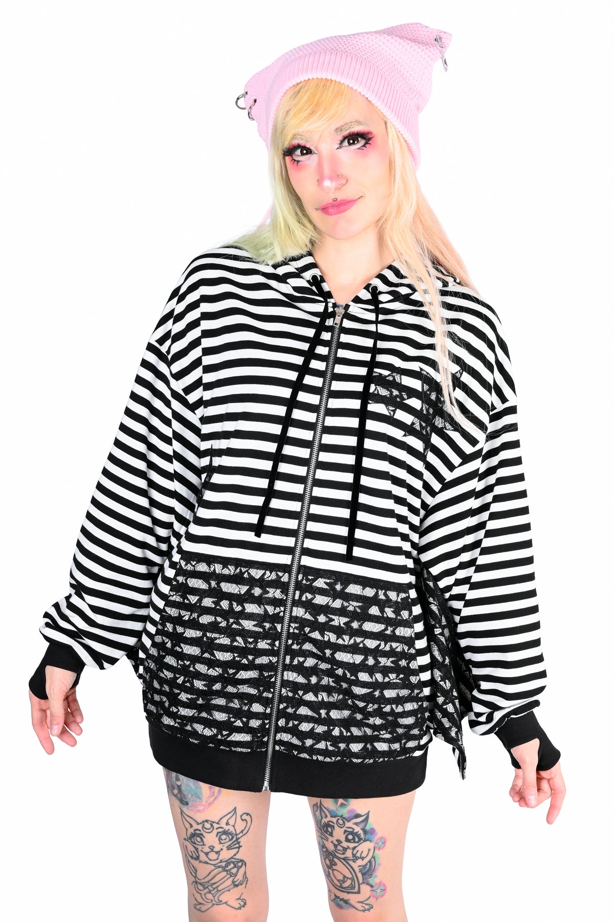 A blonde front facing model wearing a black and white striped batwing hoodie with 2 black lace star cut outs on the right chest area and one on the bottom left chest area and the same lace is on the sleeves wings and pockets, paired with a pink beret, against a solid white background.