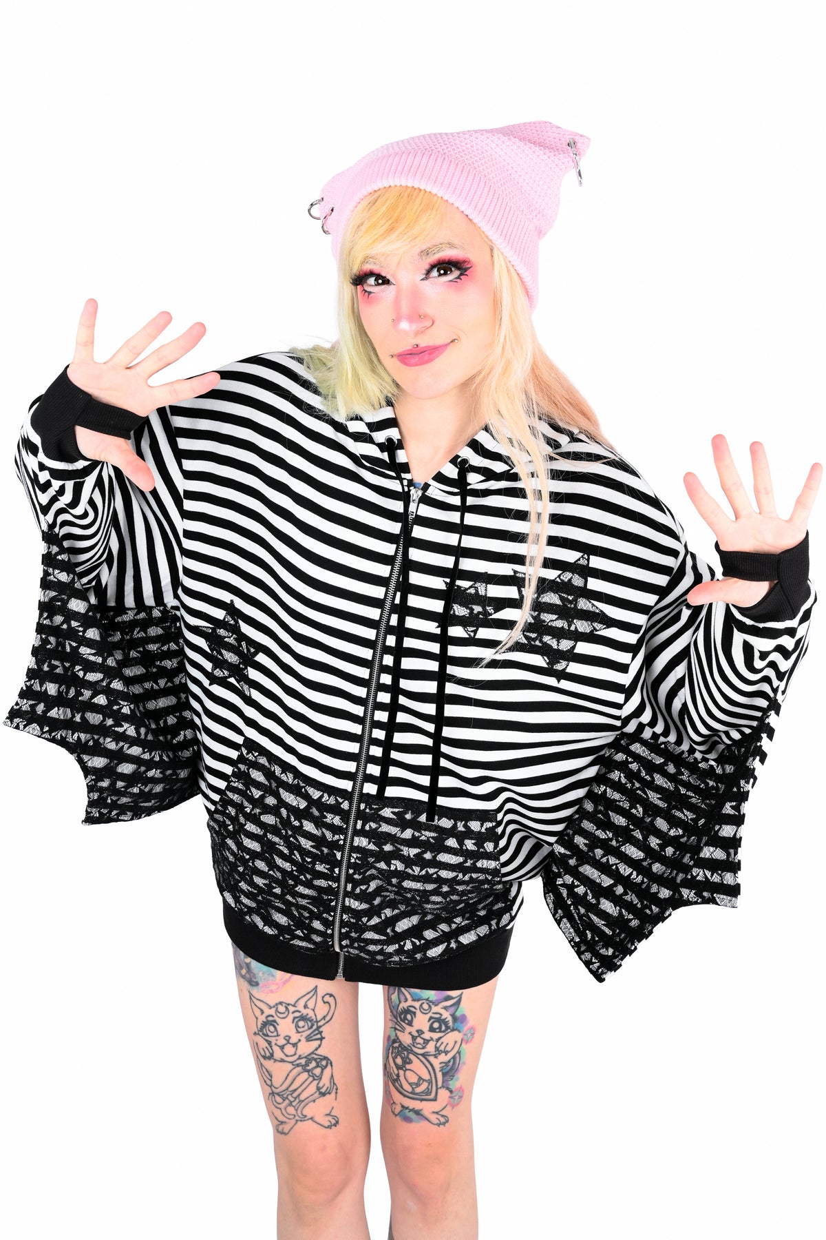 A woman model wearing a black and white striped batwing hoodie with 2 black lace star cut outs on the left chest area and 1 on the bottom right chest area. the same lace is on the sleeves wings and pockets, paired with a pink beret, against a solid white background. The model is leaning forward a little with their hands open like they are counting to 10. 