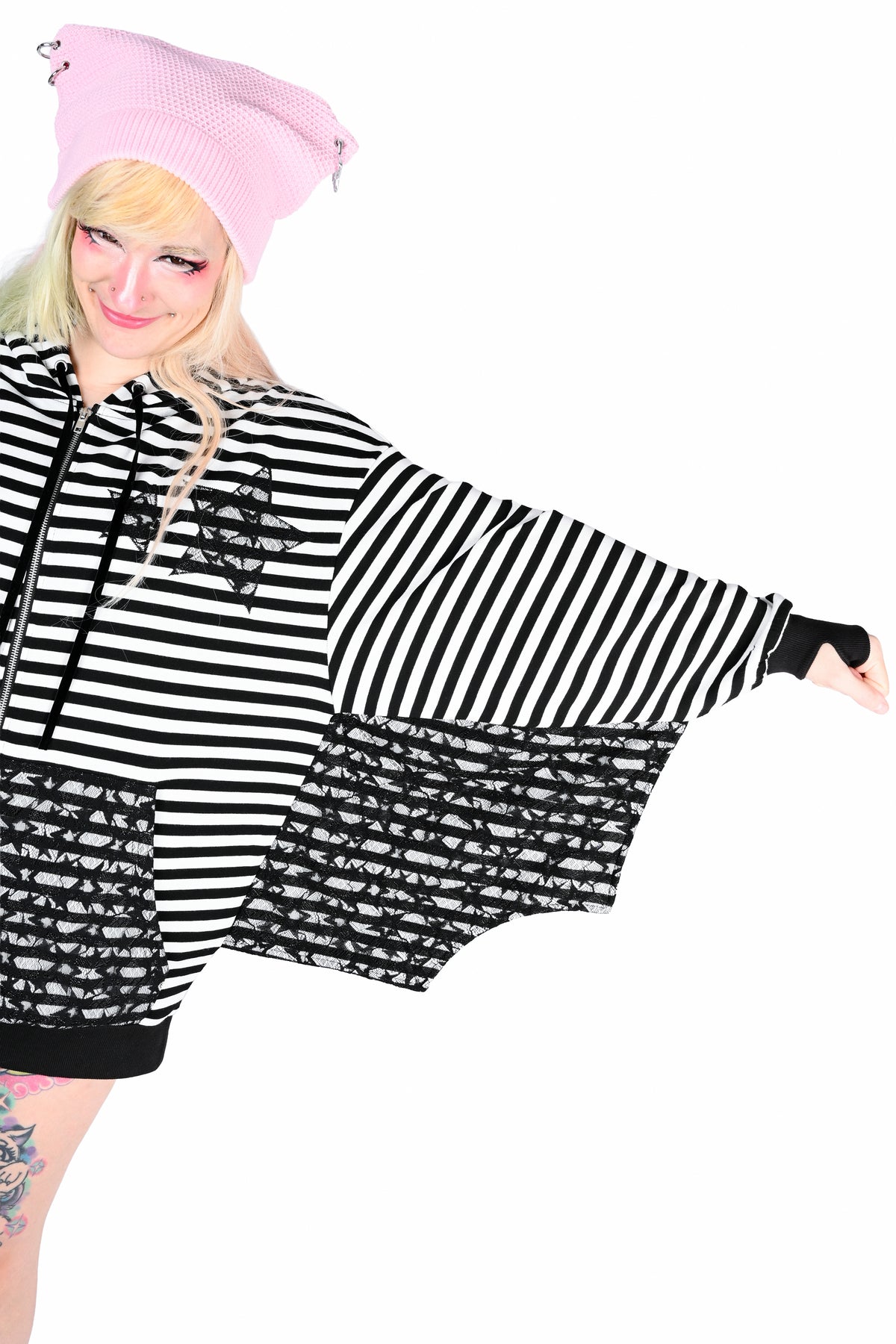 A model with a cheeky grin wearing a black and white striped batwing hoodie with 2 black lace star cut outs on the lapel and the same lace is on the sleeves wings and pockets, paired with a pink beret, against a solid white background. Her arm is outstretched to show the bat wing sleeve