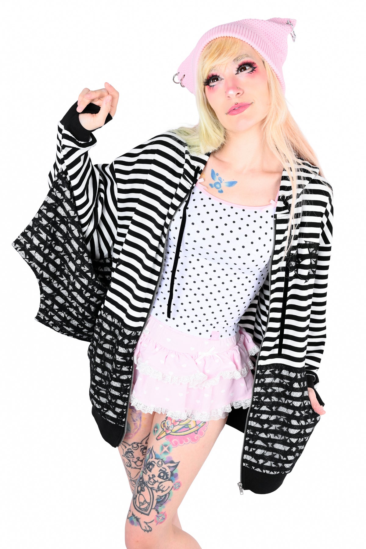 A model posing in a black and white striped zip-up hoodie with bat wings, a white polka-dot top, a light pink ruffled skirt, and a pink knit beanie, set against a plain white background. She is looking up and to the left (her right)