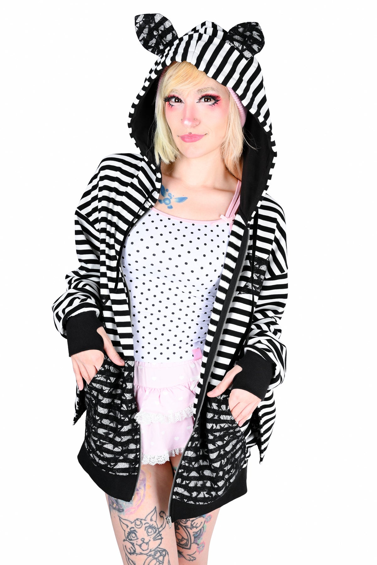 A model wearing a black and white striped hooded jacket with bat ears, a white polka dot camisole, and a light pink ruffled skirt, set against a plain white background.