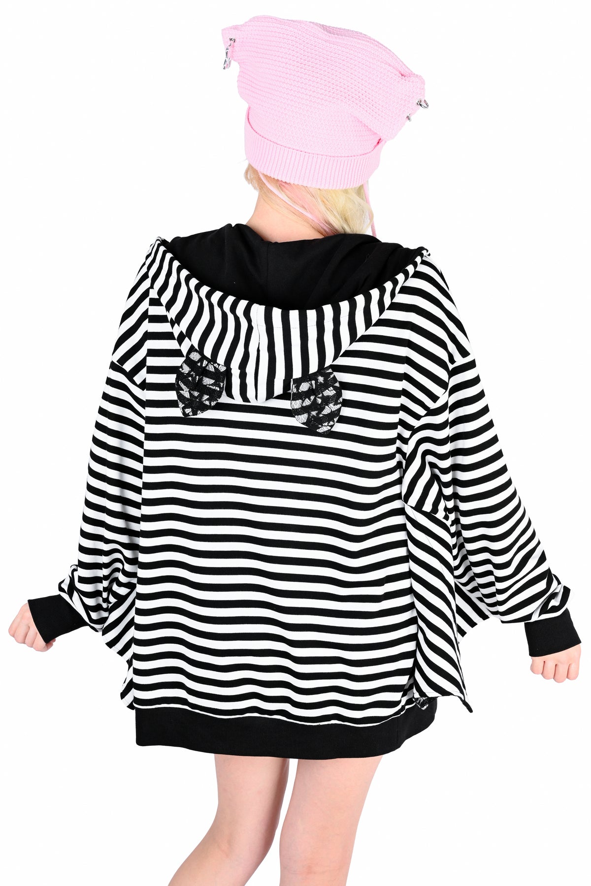 A model from behind wearing a black and white horizontally striped hoodie with cute black lace bat ears on the top of the hood. She is wearing a pink knit beanie and standing against a white background.