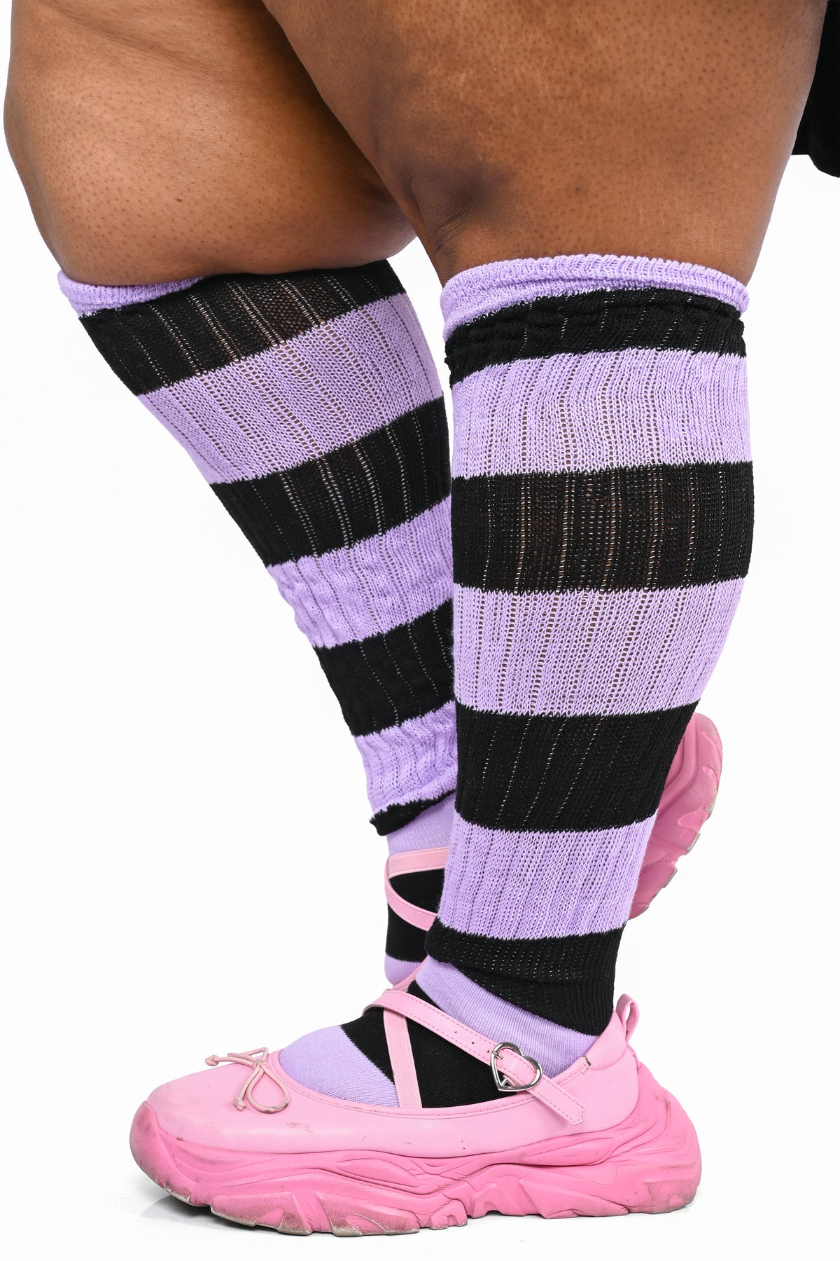 A close-up of a plus size person's legs wearing lavender and black striped ribbed knee-high socks with pink shoes against a white background.