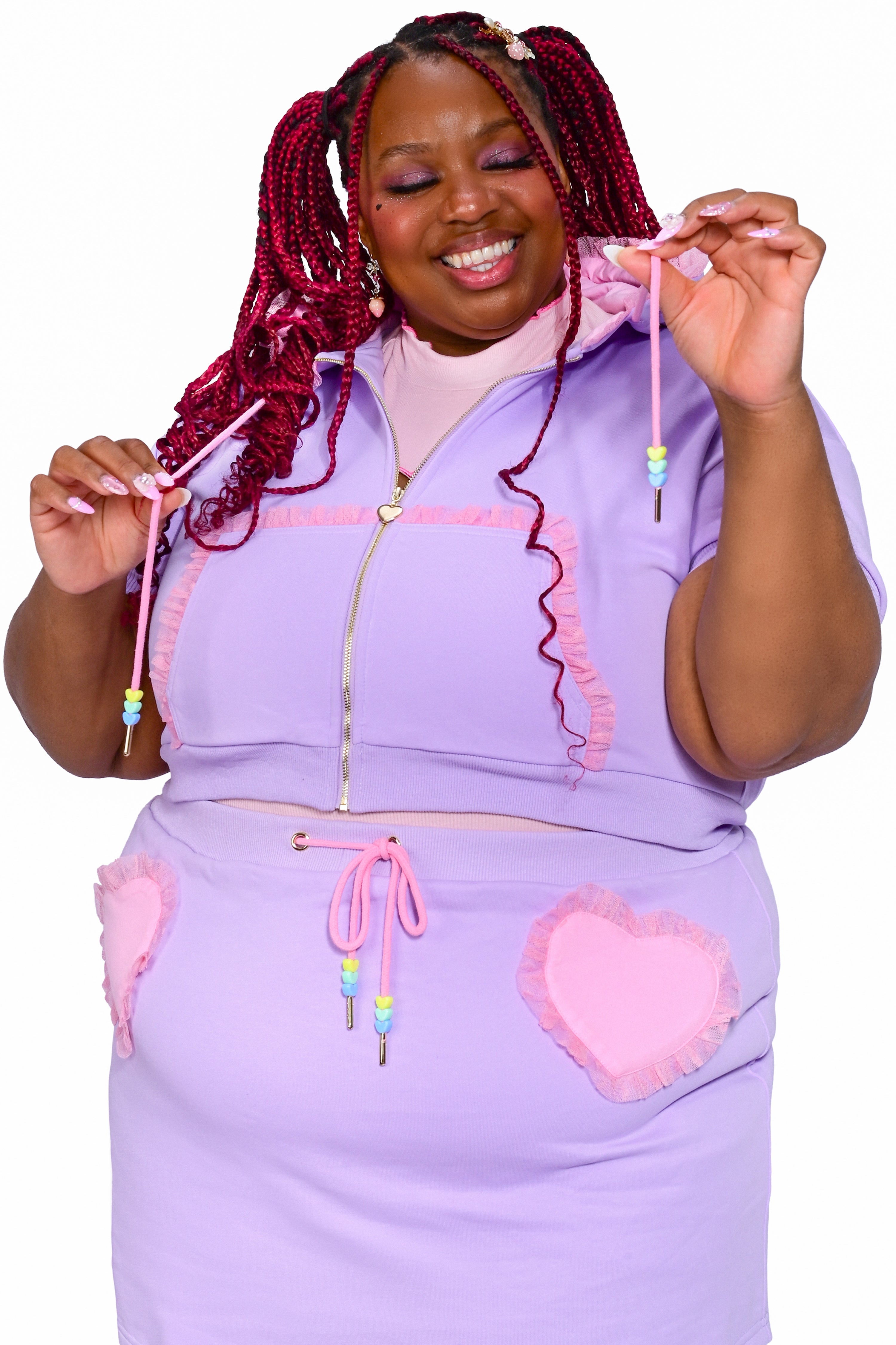 A person wearing a purple cropped hoodie with heart charms on the zipper and drawstrings, paired with a matching skirt.