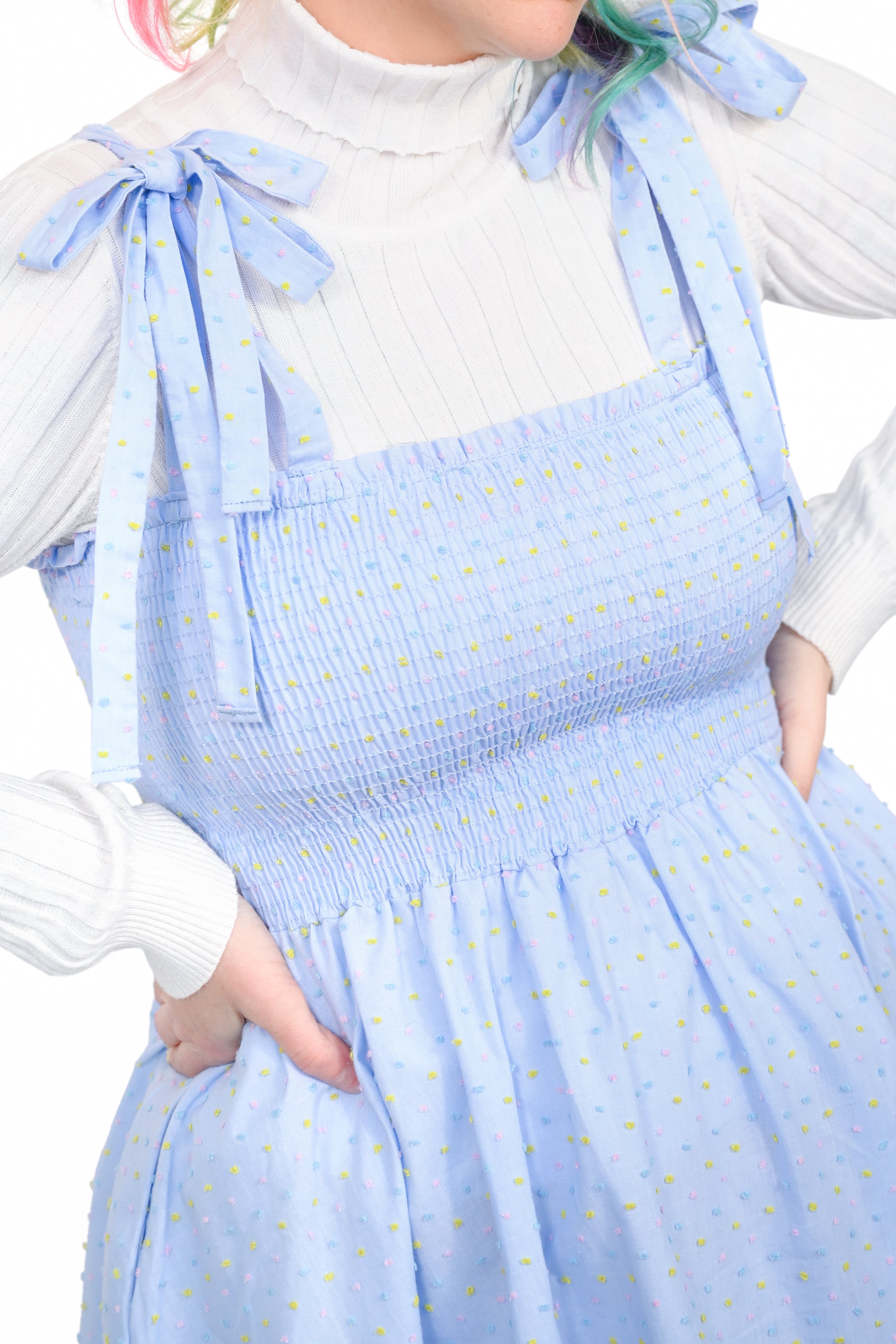 Model in a blue thigh-length sundress with a ruched bodice and tie straps. The straps are tied at the shoulders to create a  ruffled bow.